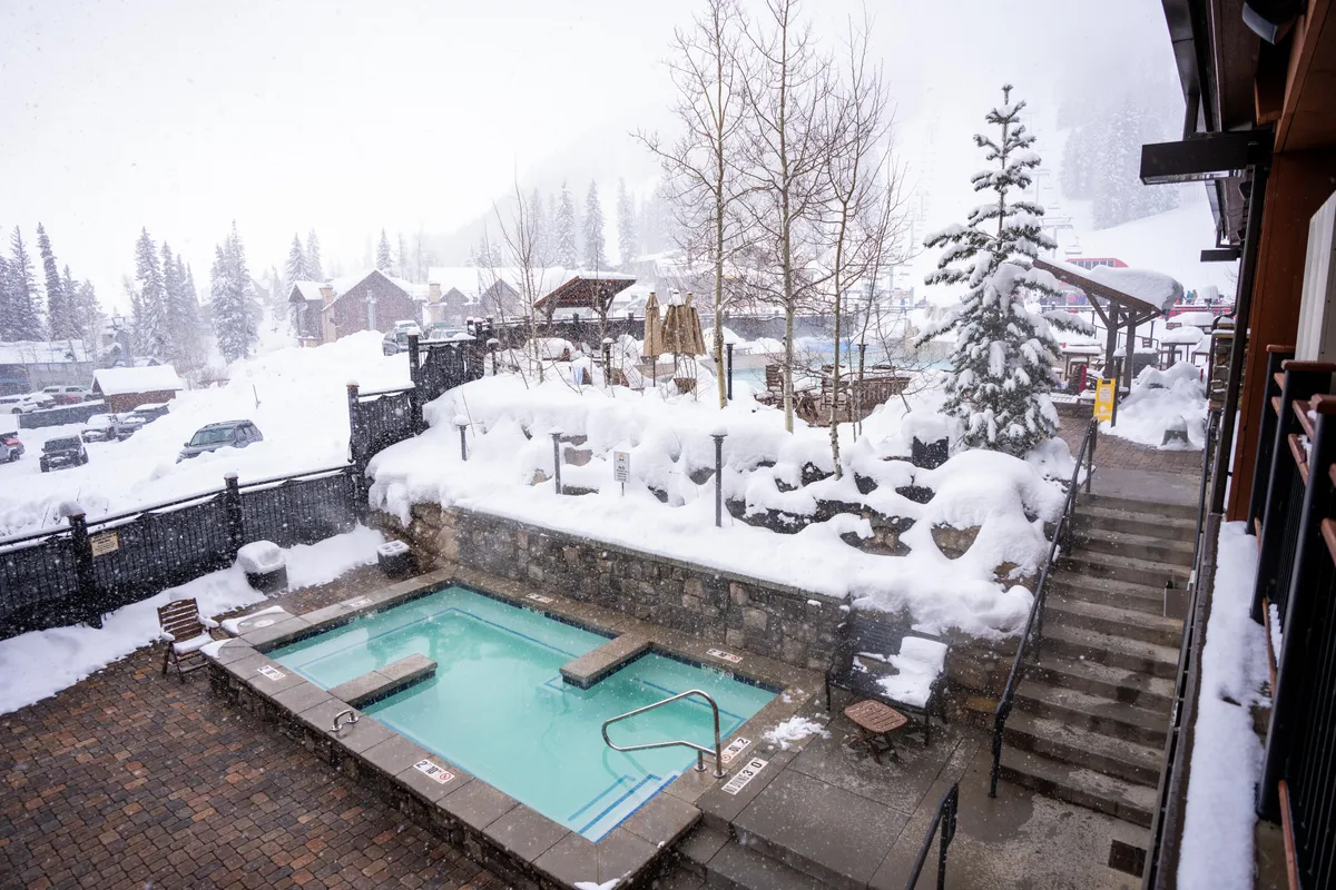 This image shows a steaming hot tub nestled in a winter wonderland, with snow-covered landscape, wooden structures, and evergreen trees. The setting is tranquil and inviting, suggesting a perfect spot to relax and unwind after a day of activities, while also emphasizing the unique experience of soaking in warmth as snow falls around.