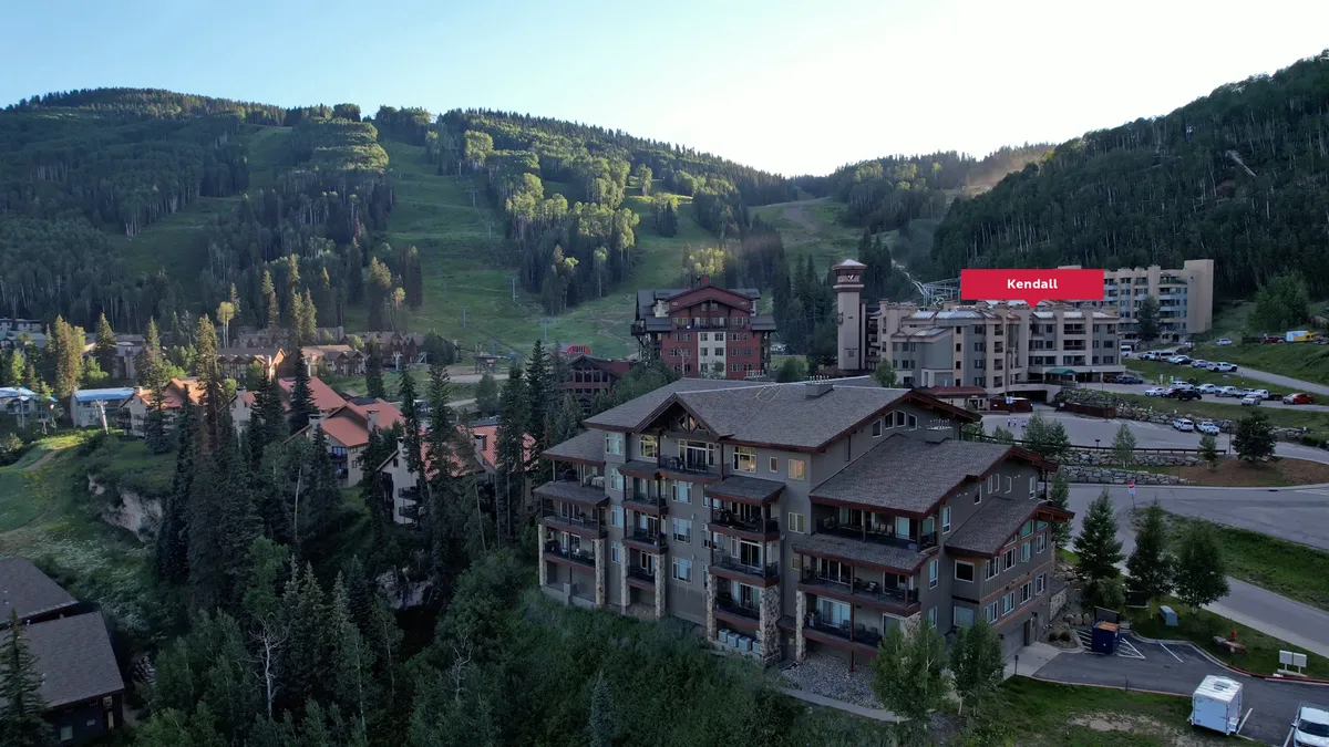 This image captures a serene mountain backdrop with residential buildings nestled among a forested area. The scene includes visible ski slopes, inviting outdoor dining setups, and hints of people exploring the village. The sunset casts a warm light over the mountains, emphasizing the tranquil yet active village atmosphere.