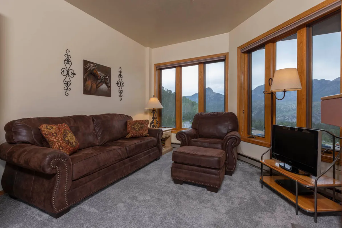 This image showcases a welcoming living area featuring a brown leather sofa, an armchair, and large windows that frame scenic mountain views. The decor is warm and inviting, with mountain-inspired wall art adding a regional touch. The room is filled with natural light, creating a cozy, tranquil setting perfect for relaxing after outdoor adventures.
