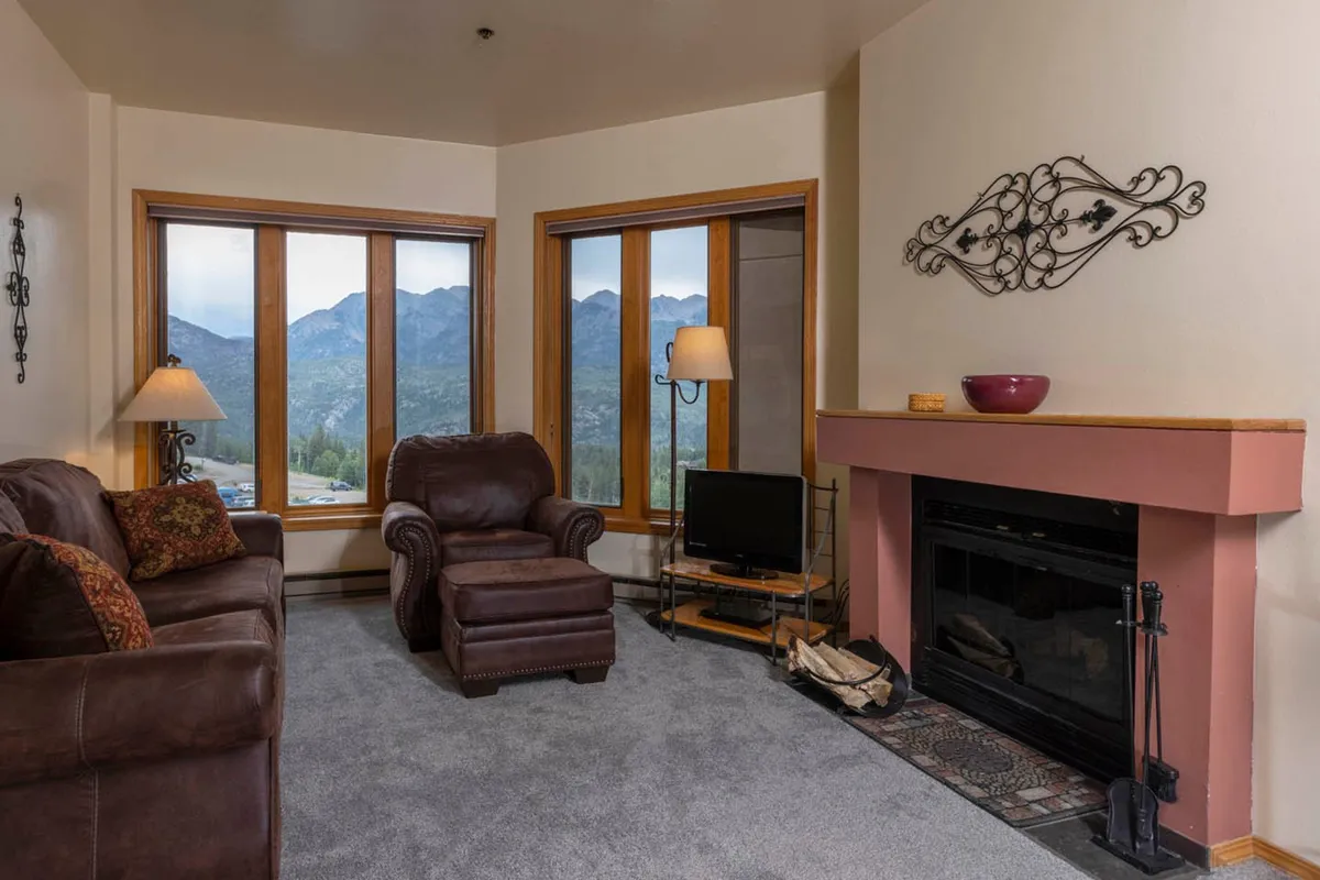 The image showcases a cozy condo interior with plush leather sofas facing a crackling wood-burning fireplace. Large windows frame breathtaking mountain views, filling the space with natural light. The inviting atmosphere is perfect for relaxing with a book, sipping coffee, or enjoying a sunset after a day of alpine adventure.