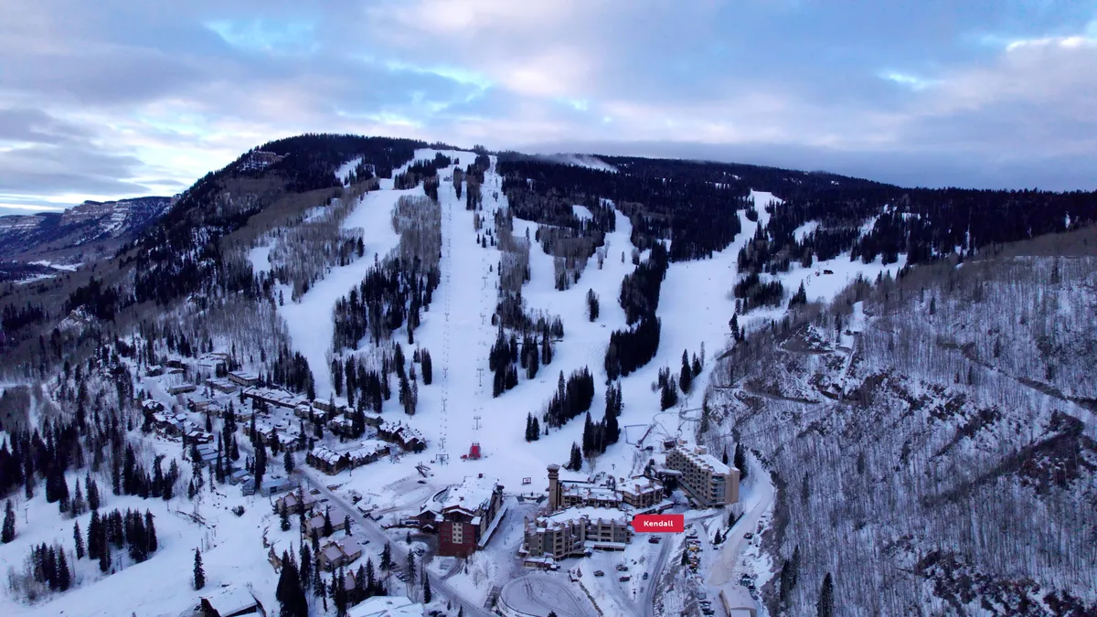 A panoramic exterior view of a lively ski resort nestled amidst a snow-blanketed mountain range. The foreground features gently sloping ski runs dotted with skiers and snowboarders, while the background reveals towering peaks and snow-covered trees. The resort structures are visible, providing a welcoming base for adventure seekers.