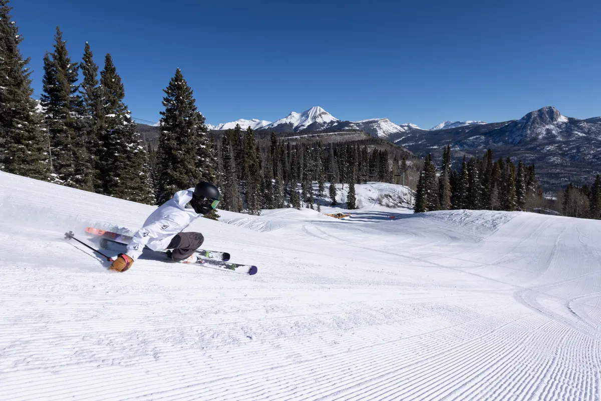 A dynamic shot of a skier carving down a pristine, snow-covered slope, framed by evergreen trees and expansive mountain views. The sense of speed and exhilaration is palpable, while other adventurers can be seen enjoying the slopes further in the distance. The image evokes the thrill and camaraderie of mountain activities.