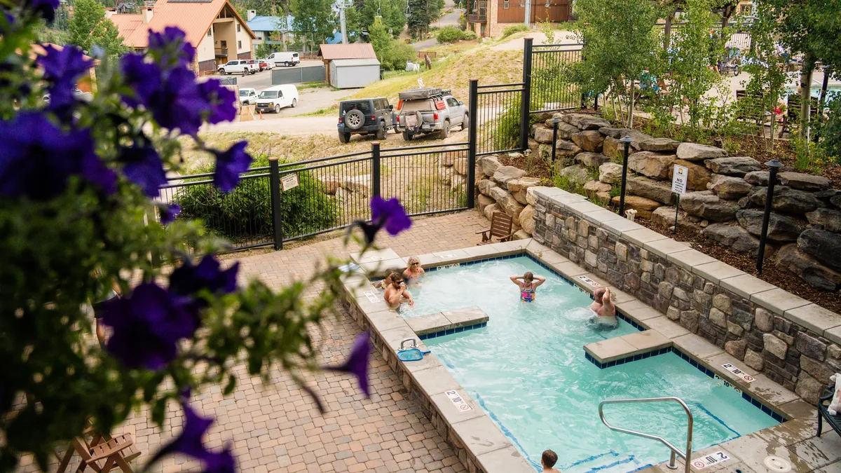 A rooftop hot tub and heated outdoor pool are featured against a backdrop of lush greenery and stone walls. The scene shows people relaxing and socializing in a laid-back, outdoor lodge environment. Poolside seating and subtle architectural details hint at the upscale amenities available to residents.
