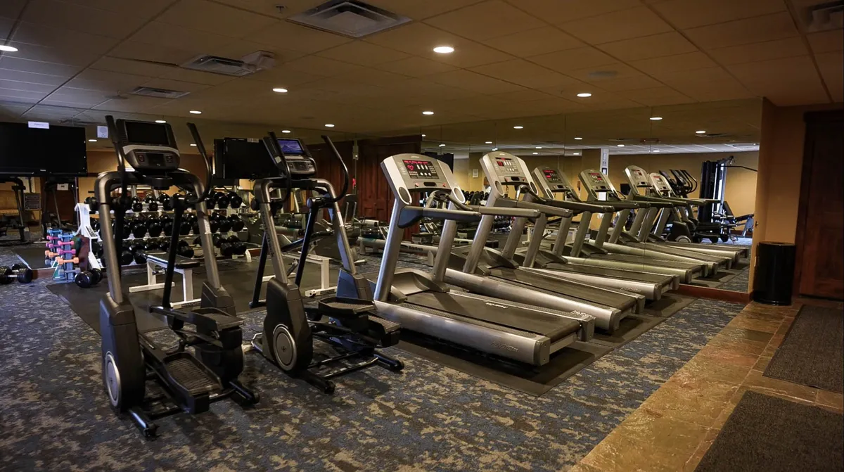 The image displays the fitness center's interior, with treadmills, ellipticals, dumbbells, mirrors, and space for group fitness. The mood is energetic and uplifting, promoting an active lifestyle and wellness routines.