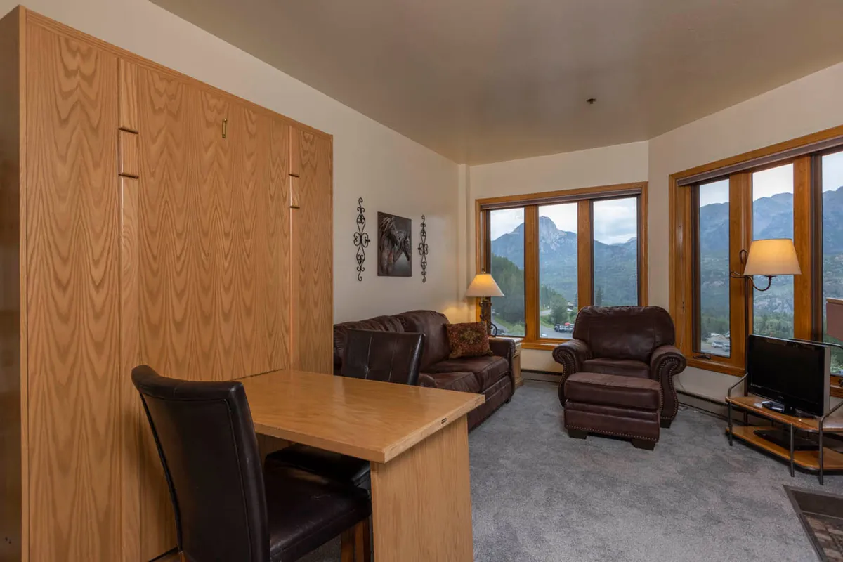 A cozy living space with large windows framing breathtaking mountain scenery. Brown leather sofas and a wooden dining table add warm, natural textures and a mountain lodge feel to the interior.