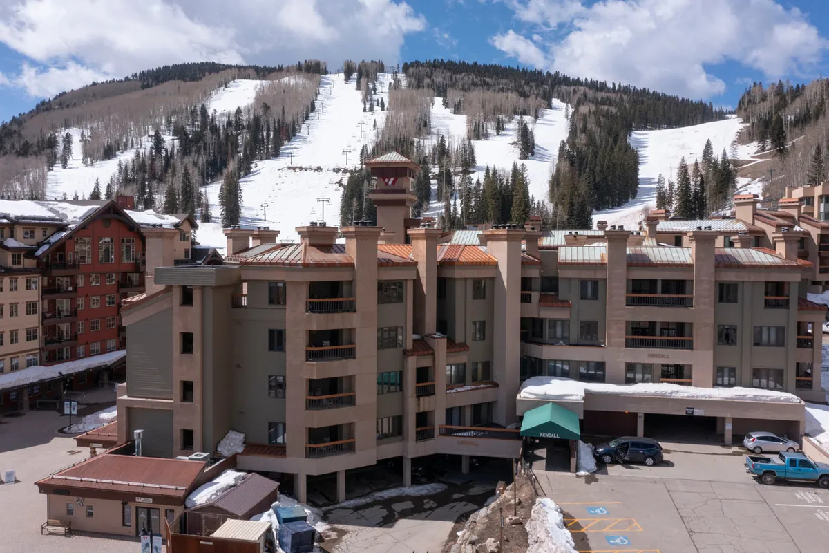 This exterior image highlights the building architecture with a clear view of the parking area in front, set against the backdrop of imposing mountain slopes. The scene captures easy access to the property from the garage or parking area, with visible pathways leading toward the village and ski lifts.